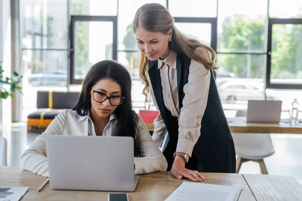 Beautiful businesswoman standing near attractive coworker in glasses and looking at laptop — Stock Photo