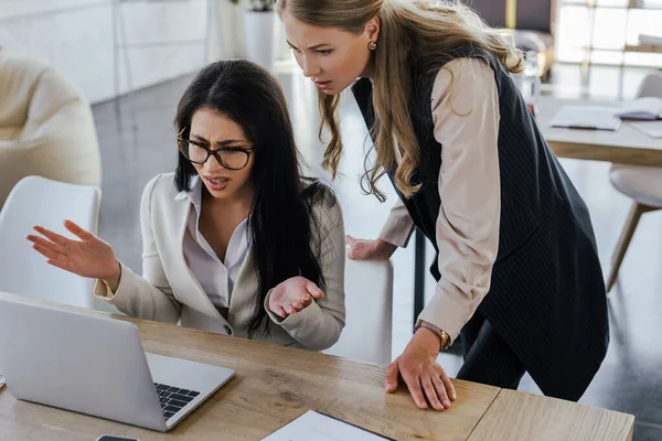 Displeased businesswoman in glasses looking at laptop and gesturing near attractive coworker — Stock Photo