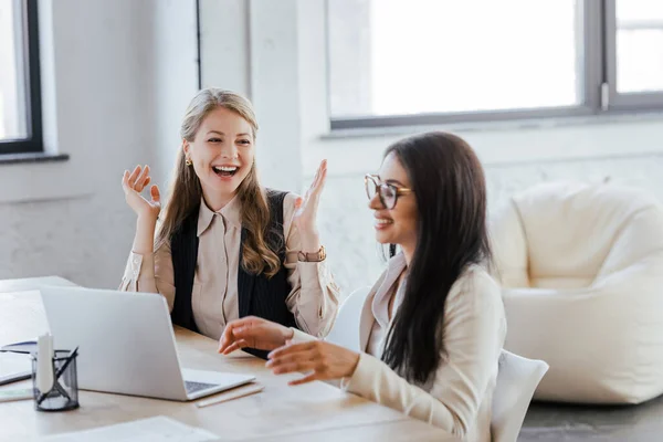 Enfoque selectivo de la mujer alegre mirando compañero de trabajo feliz cerca de la computadora portátil en la oficina - foto de stock