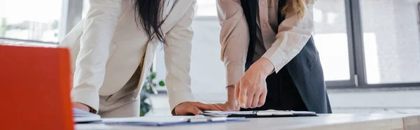 Cosecha horizontal de mujer de negocios señalando con el dedo en el portapapeles cerca de compañero de trabajo en la oficina - foto de stock