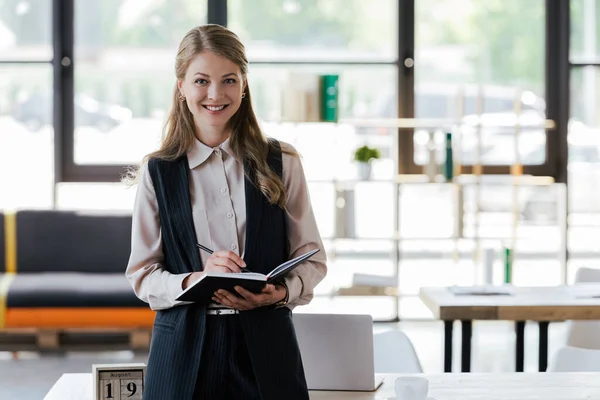 Femme d'affaires heureuse debout près de la table et tenant un carnet avec stylo dans le bureau — Photo de stock