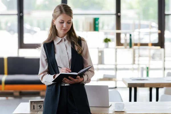 Atractiva mujer de negocios de pie cerca de la escritura de mesa en cuaderno - foto de stock