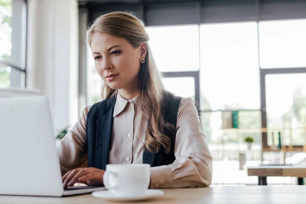 Selective focus of attractive businesswoman using laptop near cup — Stock Photo