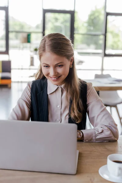 Selective focus of happy businesswoman using laptop near cup of coffee — Stock Photo