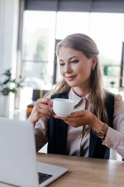 Enfoque selectivo de la mujer de negocios feliz sosteniendo la taza de café y mirando el ordenador portátil - foto de stock