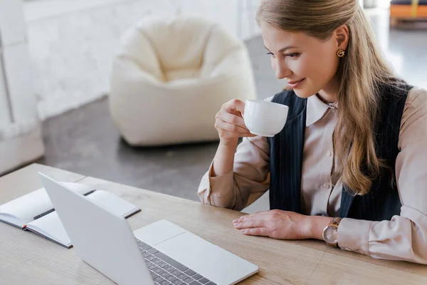 Feliz mujer de negocios sosteniendo la taza de café y mirando el ordenador portátil - foto de stock