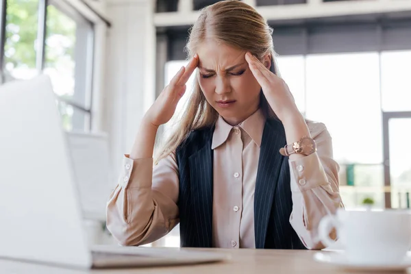 Selective focus of tired woman touching temples while suffering from migraine near laptop and cup — Stock Photo