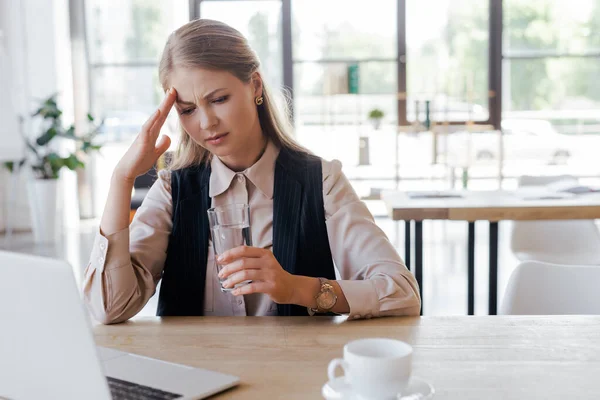 Selektiver Fokus der müden Frau, die den Kopf berührt und ein Glas Wasser in der Nähe von Laptop und Tasse hält — Stockfoto