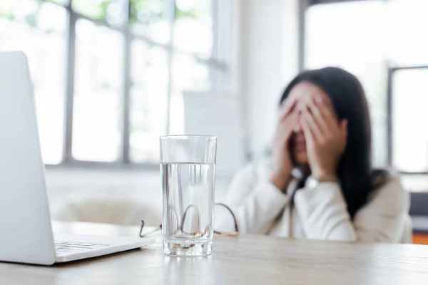 Selektiver Fokus eines Wasserglases in der Nähe von Laptop und müder Geschäftsfrau, die das Gesicht verdeckt — Stockfoto