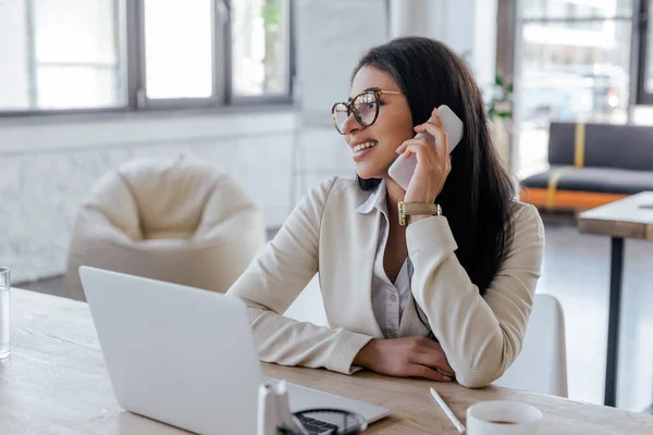 Вибірковий фокус щасливої бізнес-леді в окулярах, що розмовляють на смартфоні біля ноутбука — стокове фото