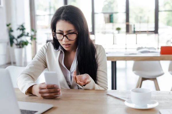 Вибірковий фокус незадоволеної бізнес-леді, використовуючи смартфон і жест біля ноутбука і чашки — стокове фото
