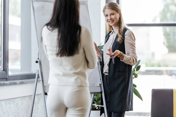 Selective focus of happy businesswoman holding marker pen near flipchart and looking at coworker — Stock Photo