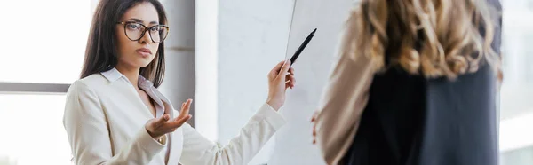 Панорамный снимок предпринимательницы в очках, держащей маркерную ручку возле флипчарта и смотрящей на коллегу — стоковое фото