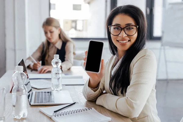 Enfoque selectivo de mujer de negocios feliz en gafas que sostienen el teléfono inteligente con pantalla en blanco - foto de stock