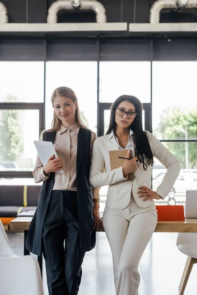 Happy businesswoman holding digital tablet near beautiful coworker in glasses standing with hand on hip — Stock Photo