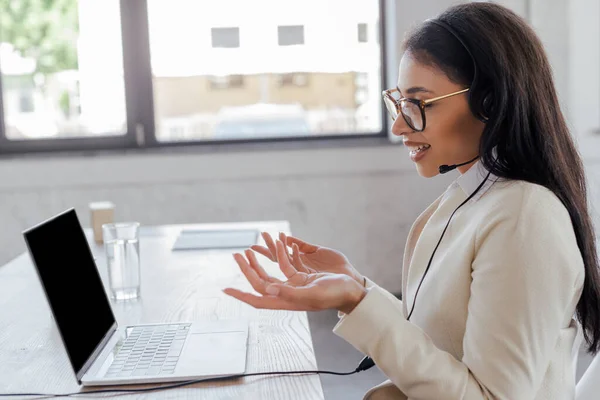 Opérateur heureux dans la gestuelle casque tout en parlant près d'un ordinateur portable avec écran vide — Photo de stock