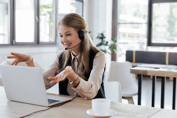 Selective focus of happy operator in headset gesturing while talking near laptop and cup — Stock Photo