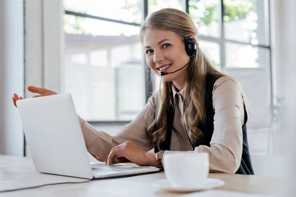 Selective focus of happy operator in headset smiling near laptop and cup — Stock Photo