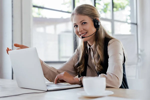 Enfoque selectivo de operador feliz en auriculares sonriendo cerca de la computadora portátil y la taza - foto de stock