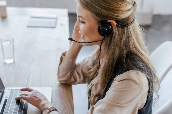 Seitenansicht des glücklichen Bedieners im Headset, der neben dem Laptop auf dem Tisch lächelt — Stockfoto