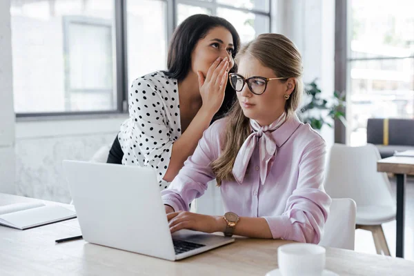 Empresária sussurrando no ouvido de colega de trabalho em óculos enquanto fofoca no escritório — Fotografia de Stock