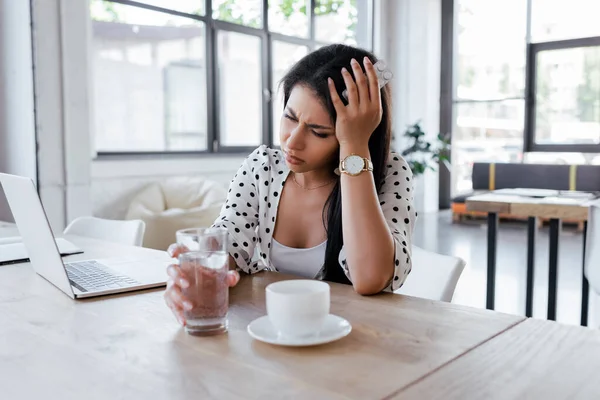 Mulher com dor de cabeça segurando vidro de água perto de xícara de café e laptop no escritório — Fotografia de Stock