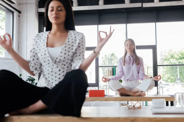 Selective focus of attractive businesswomen with closed eyes meditating on tables in office — Stock Photo