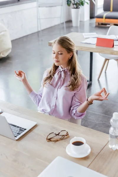 Бізнес-леді з закритими очима, роздумуючи біля столу з ноутбуком і чашкою в офісі — стокове фото