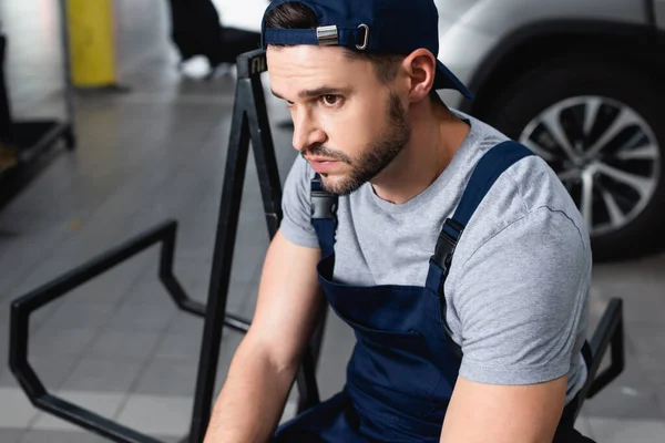 Schöner Mechaniker mit Mütze sitzt neben Auto in Werkstatt — Stockfoto