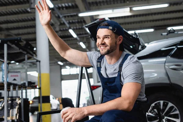 Felice meccanico in tappo agitando mano e tenendo tazza di carta in officina — Foto stock