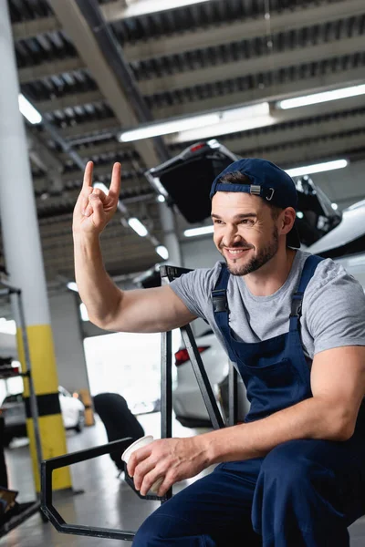 Glücklicher Mechaniker mit Mütze zeigt Felsschild und hält Pappbecher in Autowerkstatt — Stockfoto