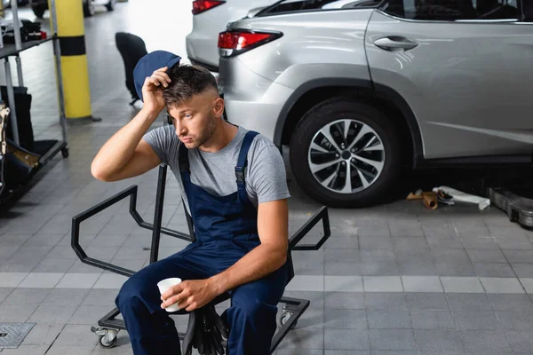 Mecánico cansado sosteniendo la tapa y la taza de papel mientras está sentado cerca de coches en el garaje — Stock Photo
