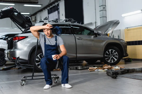 Tired technician holding cap and paper cup while sitting near cars in workshop — Stock Photo