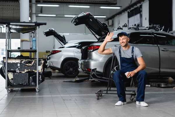 Mecánico alegre en la tapa agitando la mano y sosteniendo taza de papel cerca de los coches en el taller - foto de stock