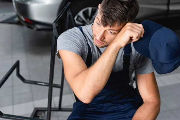 Technicien fatigué tenant chapeau et tasse en papier tout en étant assis près de la voiture dans l'atelier — Photo de stock