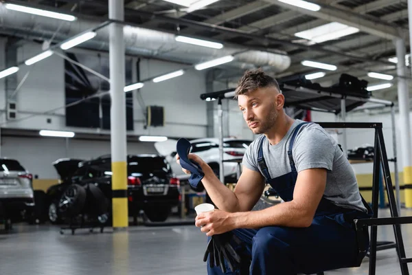 Técnico cansado em macacão segurando tampa e copo descartável enquanto sentado perto de carros na estação de serviço — Fotografia de Stock