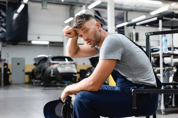 Tired technician in overalls holding cap and gloves while looking at camera and sitting near cars in workshop — Stock Photo
