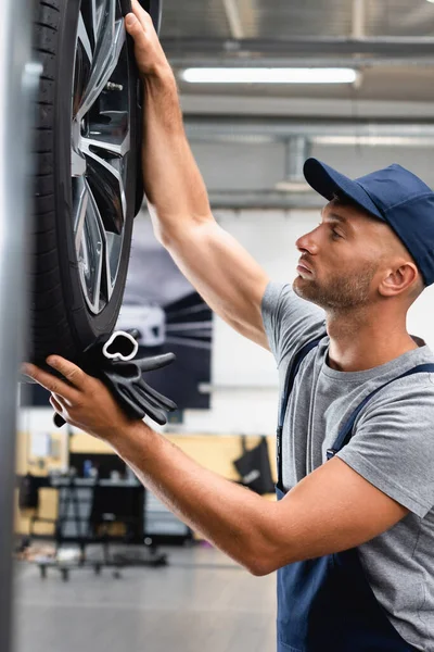 Side view of handsome technician in overalls and cap looking at car tire and holding gloves — Stock Photo