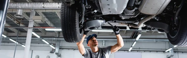 Panoramic crop of technician in cap and overalls repairing car in service station — Stock Photo