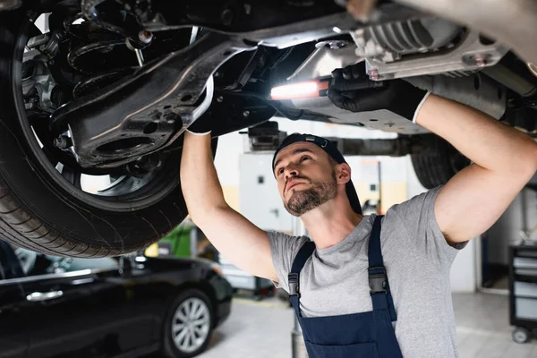 Handsome mechanic holding flashlight and fixing car in service station — Stock Photo