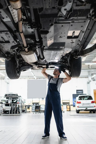 Handsome mechanic in overalls holding flashlight and repairing car in service station — Stock Photo