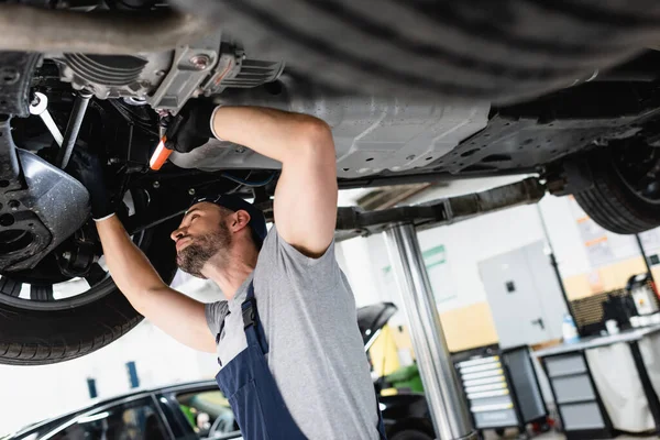 Selektiver Fokus des gut aussehenden Mechanikers in Mütze mit Taschenlampe und Schraubenschlüssel bei der Reparatur des Autos in der Tankstelle — Stockfoto