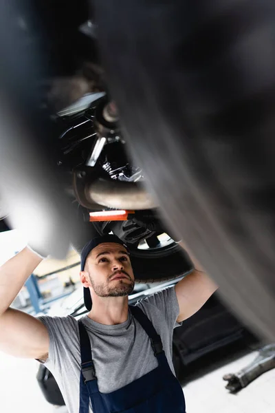 Selektiver Fokus des Mechanikers in der Mütze hält Taschenlampe während der Reparatur Auto in der Tankstelle — Stockfoto