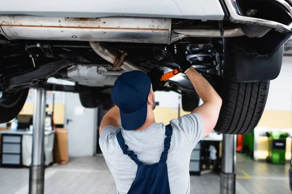 Rückansicht des Mechanikers in Mütze mit Taschenlampe und Reparatur des Autos in der Tankstelle — Stockfoto