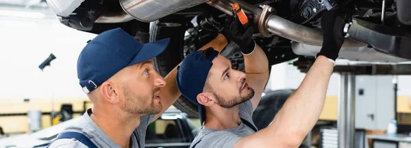 Panoramic shot of handsome mechanic in cap holding flashlight near coworker and car in service station — Stock Photo