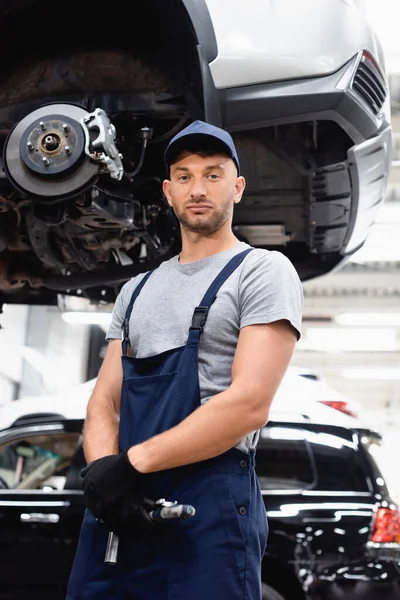 Blick des Mechanikers in Gummihandschuhen und Mütze auf die Kamera, während er in der Nähe des Autos steht — Stockfoto