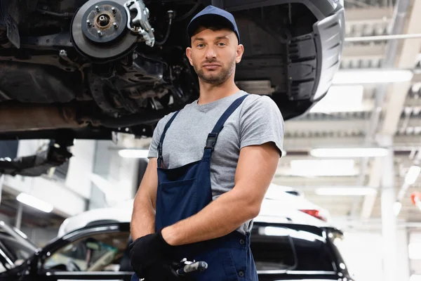 Tiefansicht des Mechanikers in Gummihandschuhen und Overalls, der in die Kamera schaut, während er in der Nähe des Autos steht — Stockfoto