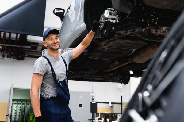 Enfoque selectivo de mecánico feliz en overoles de pie cerca del coche - foto de stock