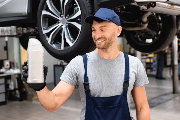 Selective focus of happy mechanic in cap and overalls holding bottle with car oil — Stock Photo