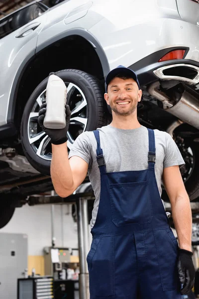 Low angle view of cheerful mechanic in cap holding bottle with car oil — Stock Photo